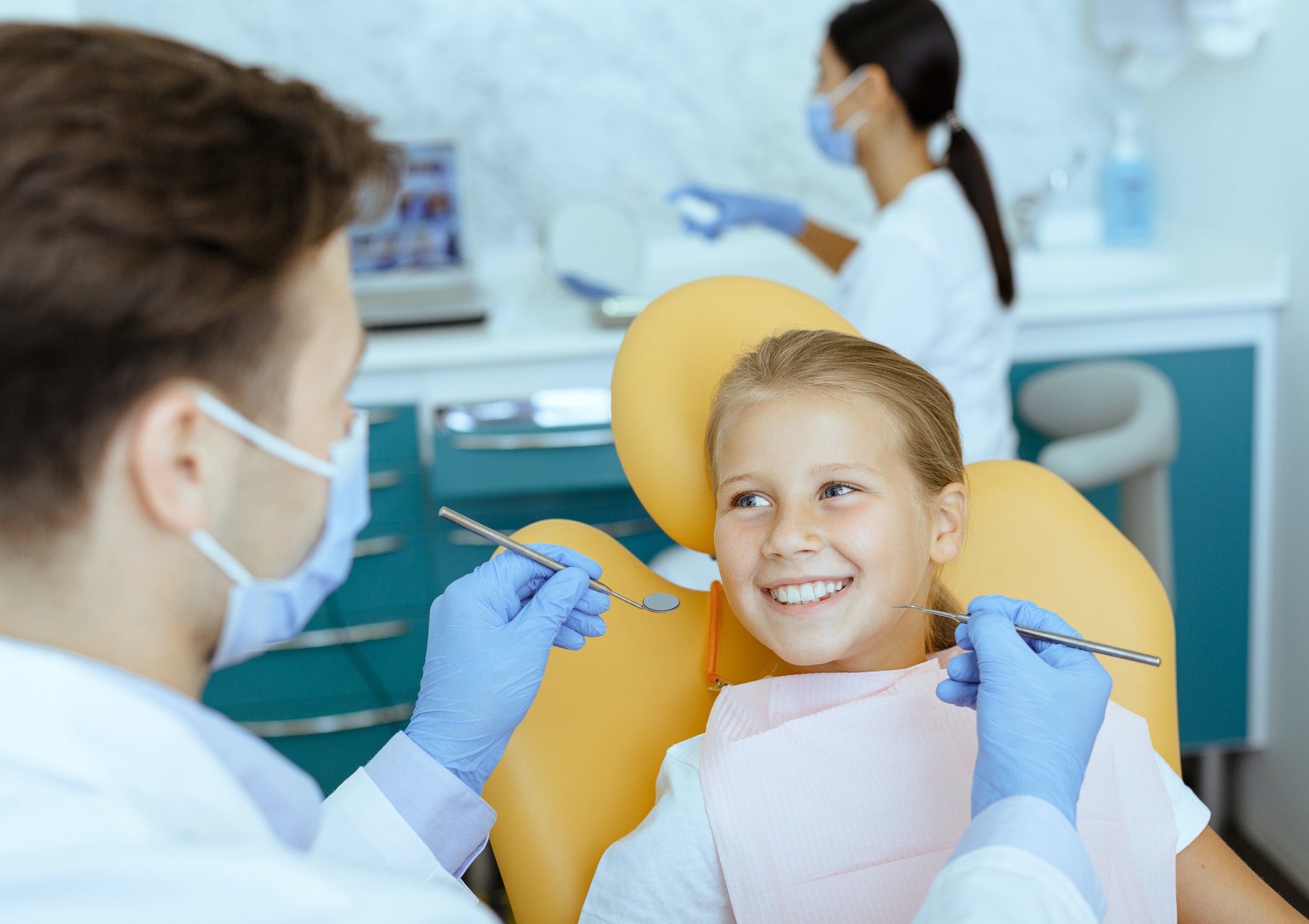 Male doctor in white coat with rubber gloves and protective mask treats teeth of smiling girl in